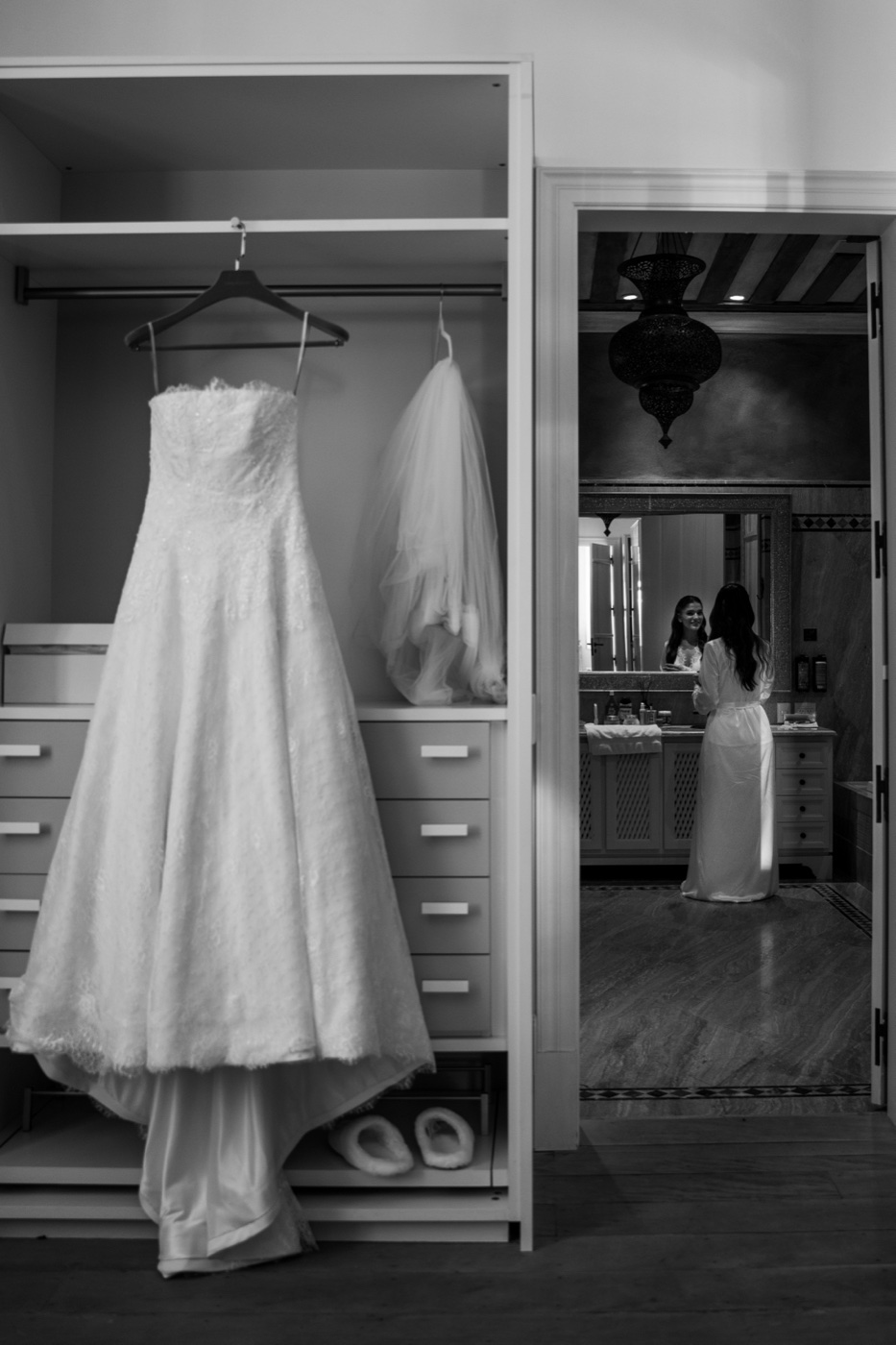 Vestido e véu pendurados no closet, com a noiva refletida ao fundo, still em preto e branco