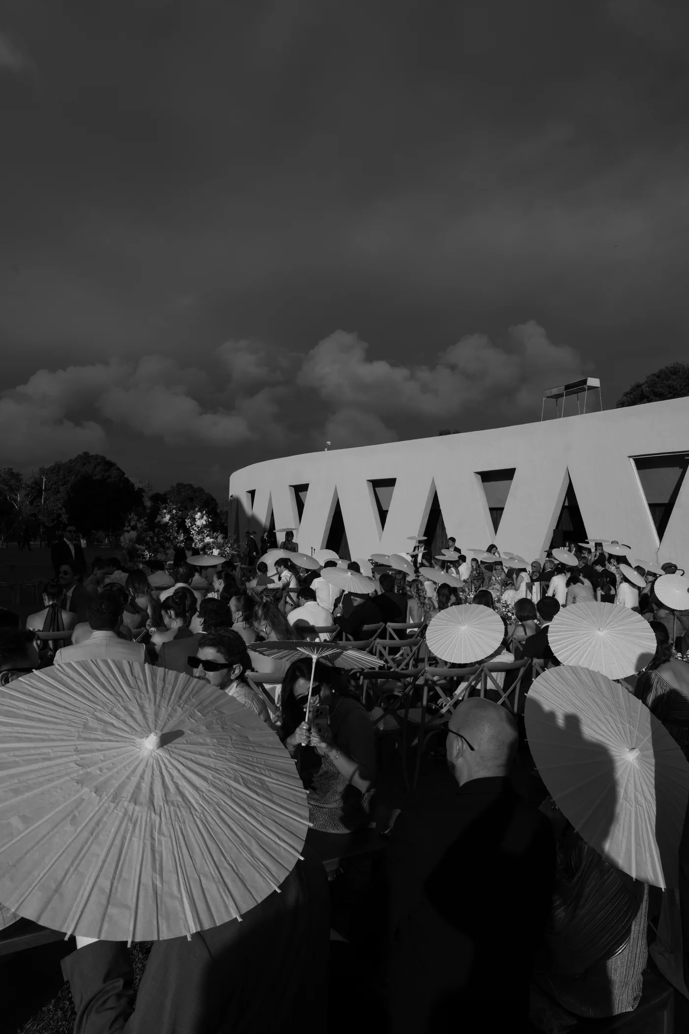 Convidados com sombrinhas brancas esperando a cerimônia, em preto e branco