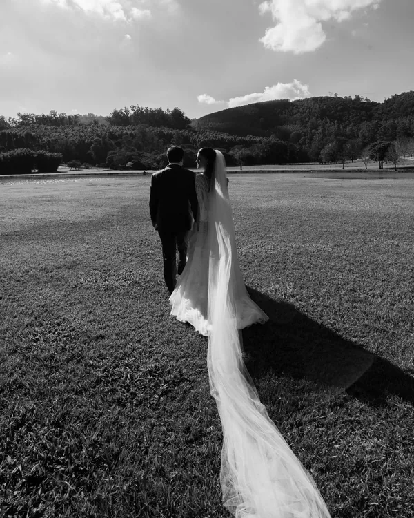 Victória e Marcos caminhando de costas no gramado, com a cauda do véu estendida atrás