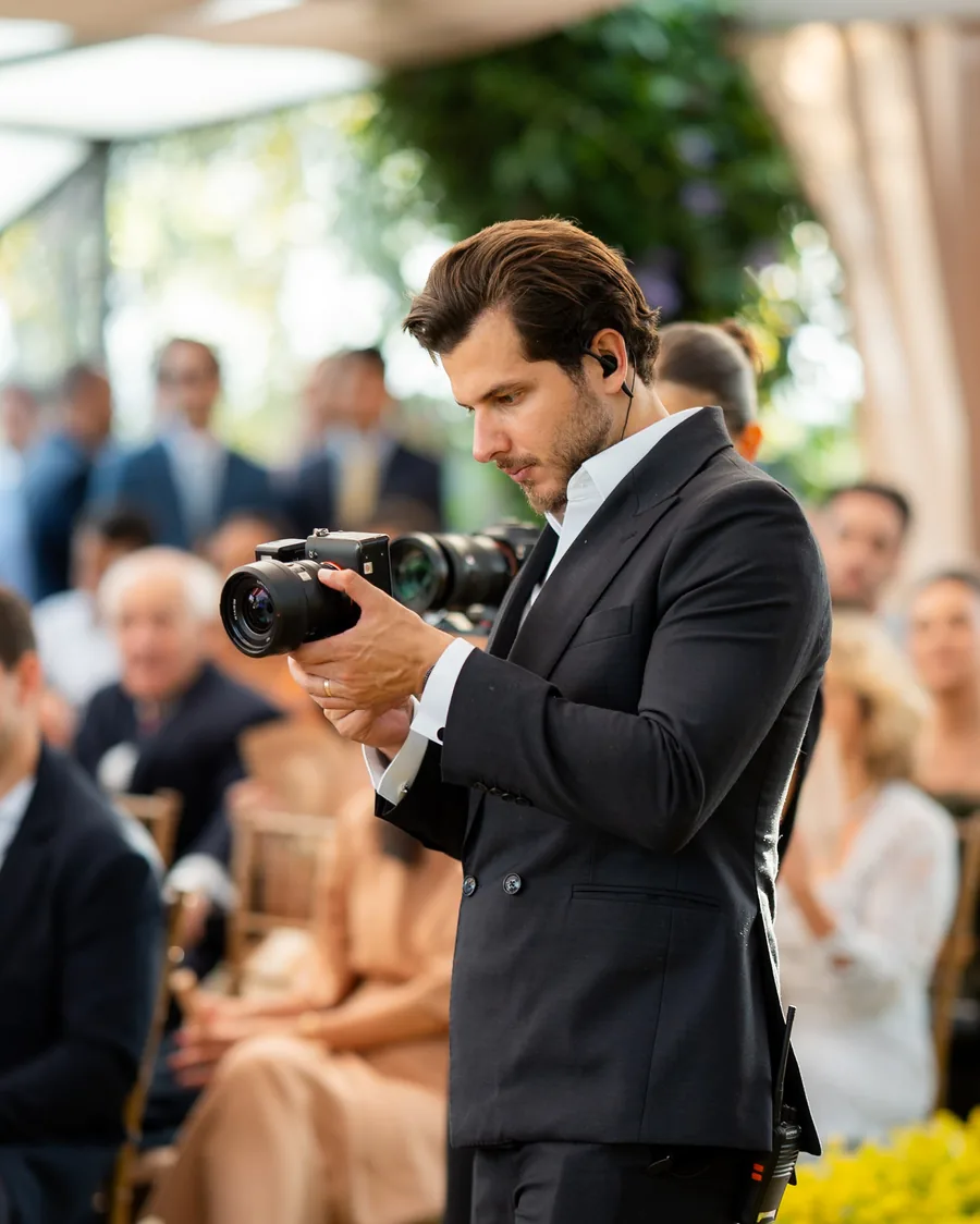 Thiago Oliveira fotografando durante uma cerimônia, de terno escuro e câmera na mão